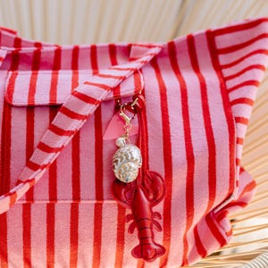 May include: A pink and red striped tote bag featuring a decorative charm attached to the handle. The charm consists of a small pink heart, a white pearl bead, a spiral seashell, and a red leather-like lobster cutout with stitched details.