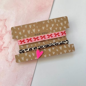 Könnte beinhalten: Drei Armbänder auf einer braunen Karte mit dem Text "FOR YOU". Das obere Armband ist rosa mit roten Schleifen, das mittlere ist im Leopardenmuster und das untere hat einen neonrosa Herzanhänger.