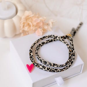 Könnte beinhalten: Ein Wickelarmband aus einem Stoffband mit schwarz-beigem Leopardenmuster. Das Armband ist mit einem kleinen, leuchtend pinkfarbenen herzförmigen Anhänger versehen, der an einem goldfarbenen Metallring befestigt ist und ein schlichtes Accessoire bildet.