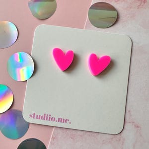 Könnte beinhalten: Ein Paar leuchtend neonpinke herzförmige Ohrstecker, die auf einer quadratischen weißen Präsentationskarte befestigt sind. Die Karte trägt den Text studiio.me., der in einer kleinen, dunkelpinken Schrift unten links aufgedruckt ist.