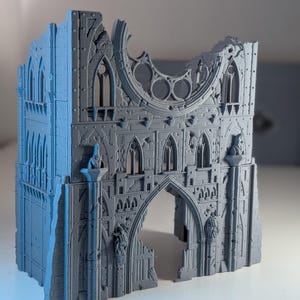 May include: A gray, gothic-style miniature building with arched doorways and windows. The model has a weathered appearance, with sections appearing broken or damaged. The structure features intricate details, including decorative elements and architectural features, suitable for tabletop gaming or model building.