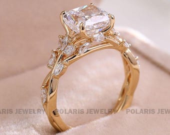 Könnte beinhalten: Ein goldener Verlobungsring mit einem großen, quadratisch geschliffenen Diamanten. Das Band hat ein florales Design mit Akzentsteinen im Marquise-Schliff. Der Ring liegt auf einer weichen, rosa Oberfläche.