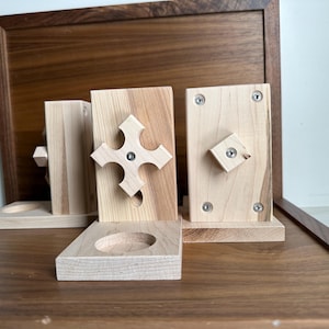 May include: A collection of wooden candle holders. One features a cross design, another a square, and a third has a geometric shape. The holders are made of light-coloured wood, set against a dark wood tray.