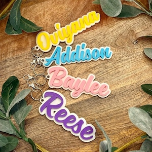May include: Four colorful name keychains with the names: Oviyana, Addison, Baylee, and Reese. Each keychain features a different color scheme, including yellow, blue, pink, and purple, with a white outline. The keychains are attached to silver key rings.