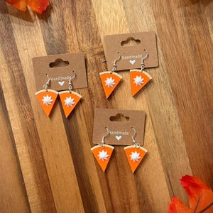 May include: Handmade pumpkin pie slice earrings with silver hooks. The earrings feature a bright orange pie filling, a white whipped cream topping, and a tan crust. The earrings are displayed on brown cardboard tags.