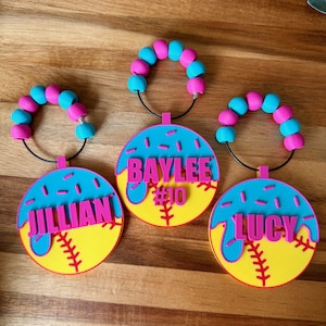 May include: Three personalized softball bag tags. Each tag features a yellow softball design with red stitching, a blue top, and a pink name. The names are Jillian, Baylee, and Lucy. Each tag has a beaded handle with pink and blue beads.