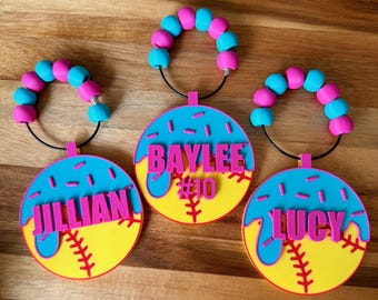 Puede incluir: Tres etiquetas personalizadas para bolsas de softbol. Cada etiqueta presenta un diseño de softbol amarillo con costuras rojas, una parte superior azul y un nombre rosa. Los nombres son Jillian, Baylee y Lucy. Cada etiqueta tiene un asa de cuentas con cuentas rosas y azules.