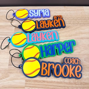 May include: A collection of colorful softball-themed name tags. Each tag features a yellow softball design with red stitching and a name or title in bold, vibrant colors. Names include "Syria", "Layken", "Harper", and "Brooke".