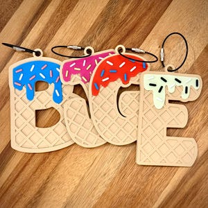 May include: Four luggage tags spelling out the word "BABE". Each tag is shaped like a waffle cone with colorful ice cream and sprinkles. The ice cream colors are blue, pink, red, and green. Each tag has a black loop for attachment.