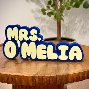 May include: A blue and yellow wooden sign that reads "MRS. O'MELIA" in a playful, rounded font. The sign is placed on a wooden table, with a small potted plant in the background. The letters are yellow with a blue outline.