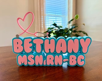 Puede incluir: Un letrero de escritorio turquesa y rosa con el nombre "Bethany" en letras de burbujas, con "MSN, RN-BC" debajo. Un estetoscopio rosa en forma de corazón se encuentra encima del letrero. El letrero está sobre una superficie de madera.