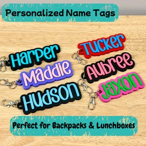 May include: Colorful personalized name tags in various colors, including blue, pink, red, and green. Each tag features a name in a contrasting color with a black outline. The tags are attached to silver keychains. The text at the top says "Personalized Name Tags".