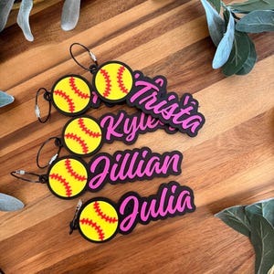 May include: Four personalized softball bag tags with names in pink script. Each tag features a yellow softball design with red stitching and a black border. The names are Trista, Kyle, Jillian, and Julia.