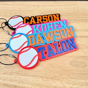 May include: Colorful baseball-themed keychains with names: Carson, Kohen, Dawson, and Talon. Each keychain features a baseball design with red stitching and a name in bold, contrasting colors. The keychains are attached to a metal loop.