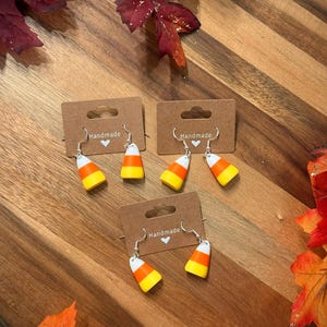 May include: Handmade candy corn earrings displayed on brown cards. The earrings feature a white, yellow, and orange design, with silver-colored hooks. The cards are on a wooden surface, surrounded by autumn leaves.