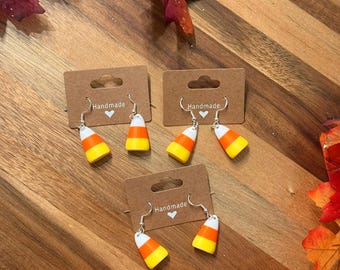 May include: Handmade candy corn earrings displayed on brown cards. The earrings feature a white, yellow, and orange design, with silver-colored hooks. The cards are on a wooden surface, surrounded by autumn leaves.
