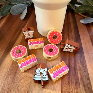 May include: A collection of colorful shoe charms featuring donuts, iced coffee, and the text "Dunkie Junkie." The charms are in shades of pink, brown, and white, and are arranged on a wooden surface near a white mug.