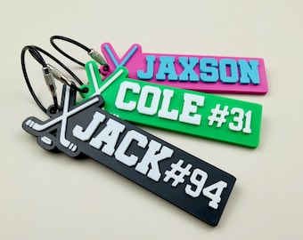 May include: Three rectangular silicone bag tags with hockey stick icons and metal cable loops. The tags are black with white text JACK #94, green with white text COLE #31, and pink with light blue text JAXSON. Each tag features a raised hockey graphic.