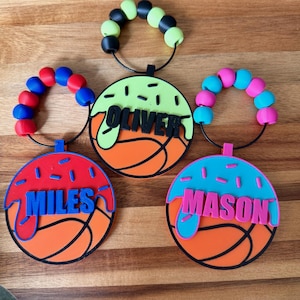 May include: Three basketball-themed name tags with colorful accents. Each tag features a basketball design with a name in bold letters: "MILES", "OLIVER", and "MASON". The tags have a beaded loop in red/blue, black/green, and pink/blue.