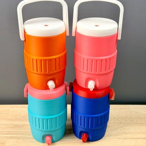 May include: Four colorful beverage coolers in orange, pink, blue, and red, each with a white lid and handle. The coolers have a spigot for dispensing drinks. The coolers are stacked in pairs, with a smaller cooler on top of a larger one.