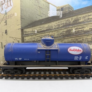 May include: A blue model train tank car featuring the True Value logo in a white oval on the side. The car has a central dome, a small ladder, and black undercarriage details, positioned on a section of model train track with black ties.