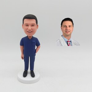 May include: A bobblehead figurine of a person in a blue medical uniform, with a smiling face. The figurine is standing on a white base. A photo of a man in a white coat is to the right of the figurine.