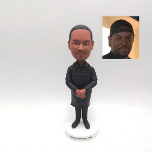 May include: A bobblehead figurine of a chef in a black uniform, with arms crossed. The figurine is on a white base with the text "happy birthday chef". A small photo of a man wearing a black cap is in the upper right corner.