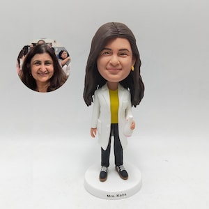 May include: A custom bobblehead figurine of a woman wearing a white lab coat over a yellow shirt and black pants. The figurine is holding a white object and stands on a white base with the name "Mrs. Kalra" printed on it. A photo of the woman is in the background.