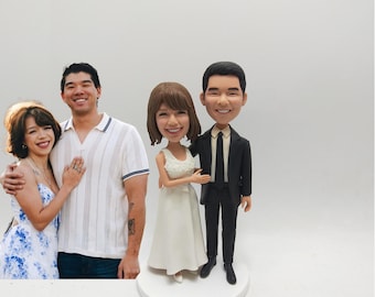 May include: A wedding cake topper featuring a bride and groom bobblehead figurine. The bride wears a white dress, and the groom is in a black suit. A couple stands next to the figurine, the woman in a blue floral dress and the man in a white polo shirt.