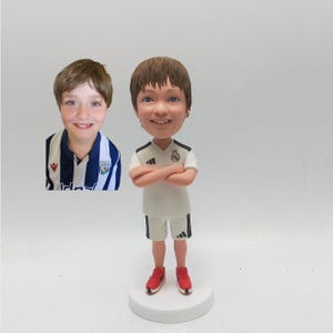 May include: A bobblehead figurine of a young person in a white football kit with black detailing, red trainers, and crossed arms. A photo of a young person in a blue and white striped jersey is to the left of the figurine. The figurine is on a white base.