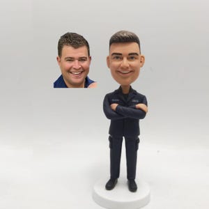 May include: A navy blue bobblehead figurine of a man in a uniform with crossed arms, standing on a white base. The figurine has a smiling face and is wearing a navy blue uniform. A photo of a smiling man is in the background.