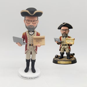 Può includere: Due statuette a testa oscillante di un uomo in abito in stile coloniale, con un computer portatile e una pergamena. La figura più grande è su una base bianca con la scritta "Chancellor of the systems". La pergamena recita "All deliverables shall be accounted for."