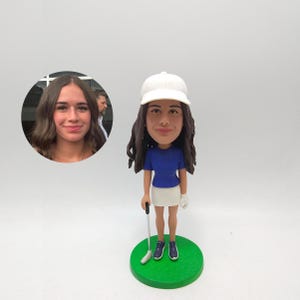 Puede incluir: Figura de cabeza oscilante de una golfista con gorra blanca, camisa azul y falda blanca. La figura sostiene un palo de golf y está de pie sobre una base verde. Una foto de una mujer está en el fondo.