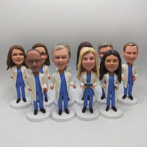 May include: A collection of bobblehead figurines depicting a medical team. Each figure wears a white lab coat over a blue uniform. The figurines stand on white circular bases, with varying skin tones and hairstyles.