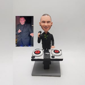 May include: A DJ figurine in a black suit, holding a microphone, with a smiling face. The figurine is positioned behind a miniature DJ setup with two turntables and a mixer. The base is grey and has the text "DJ Vincent Vega".
