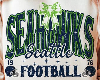 Könnte beinhalten: Ein cremefarbenes Sweatshirt mit dem Text "SEAHAWKS" in Grün und Marineblau, mit einer grünen Schleifengrafik. Unter dem Teamnamen steht das Wort "Seattle" in Schreibschrift, zusammen mit den Zahlen "19" und "76" und dem Wort "FOOTBALL".