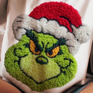 May include: A cream-colored sweatshirt featuring a green Grinch face wearing a red and white Santa hat. The Grinch's face has yellow eyes and a mischievous grin. The sweatshirt is a cozy, festive garment.