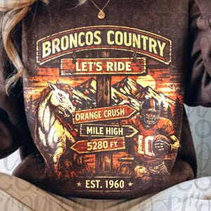 May include: Brown sweatshirt with a graphic featuring a horse, football player, and the text "Broncos Country Let's Ride." The design includes signs pointing to "Orange Crush" and "Mile High 5280 FT." The sweatshirt also has "EST. 1960" printed on it.