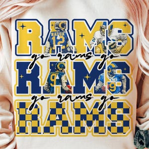 May include: A beige sweatshirt with a blue and yellow graphic that says "RAMS" in large letters. The graphic includes images of football players and the words "go rams go".