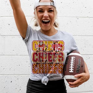Peut inclure: T-shirt gris avec l'inscription "CHIEFS" en rouge et or, avec des images de joueurs. Un ballon de football Wilson marron et blanc est tenu dans la main. Le t-shirt est nou&eacute; &agrave; la taille.