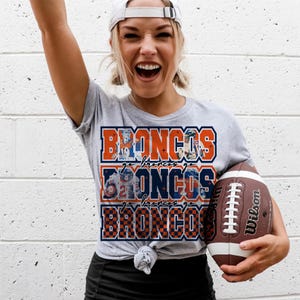 May include: A gray t-shirt with the word "BRONCOS" in orange and navy blue, stacked three times. The shirt is paired with a white baseball cap and a brown and white football.