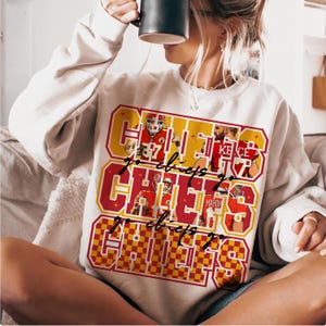 Peut inclure: Sweat-shirt cr&egrave;me avec un motif graphique avec le texte "Go Chiefs" en rouge et jaune, avec des images de joueurs de football. Le design a un style r&eacute;tro et coll&eacute;gial.