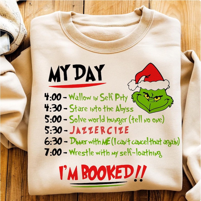 May include: A beige sweatshirt with the text "MY DAY" in black above a schedule of activities, including "Wallow In Self Pity" and "Solve world hunger." A green Grinch face with a Santa hat is on the right. The words "I'M BOOKED!!" are at the bottom.
