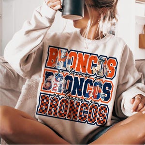 May include: Cream-colored sweatshirt featuring the word "BRONCOS" in a layered design with orange and blue accents. The design includes images of football players and the phrase "Go Broncos" repeated. The sweatshirt has a crew neck.