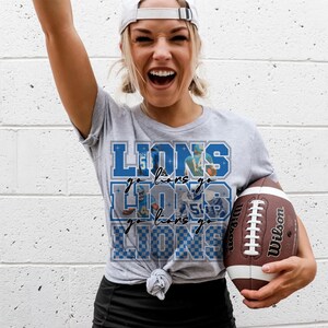 May include: A light gray t-shirt with the word "LIONS" in blue, along with the phrase "go lions go" and images of football players. The person is holding a brown and white Wilson football.
