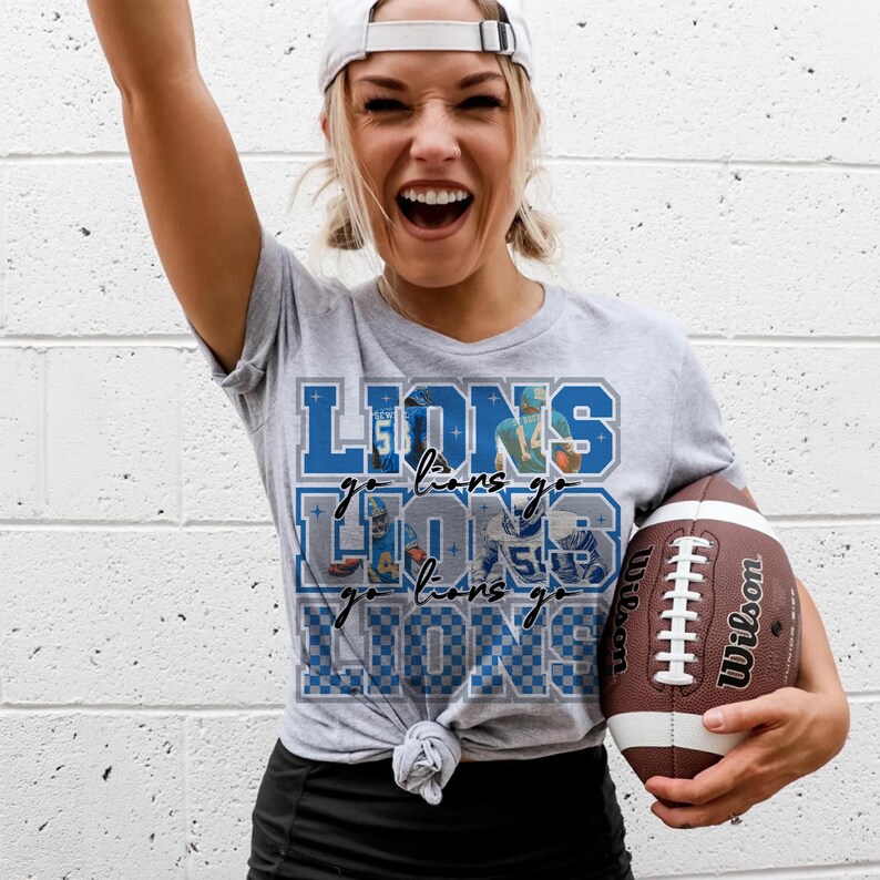 May include: A light gray t-shirt with the word "LIONS" in blue, along with the phrase "go lions go" and images of football players. The person is holding a brown and white Wilson football.