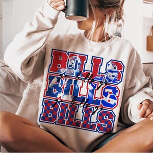 May include: Cream-colored sweatshirt with a red, white, and blue graphic that says "BILLS" multiple times. The graphic includes images of football players and helmets. The sweatshirt has a crew neck and long sleeves.