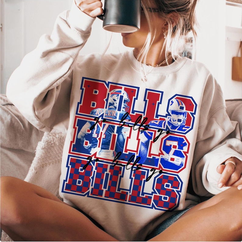 May include: Cream-colored sweatshirt with a red, white, and blue graphic that says "BILLS" multiple times. The graphic includes images of football players and helmets. The sweatshirt has a crew neck and long sleeves.