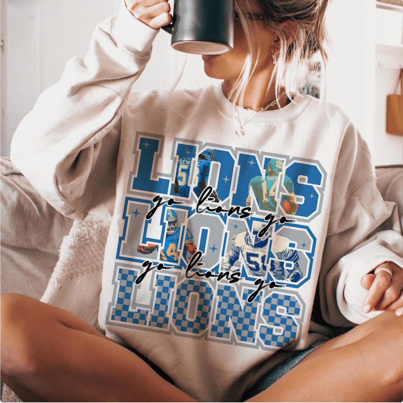 May include: A cream-colored sweatshirt featuring the text "LIONS" in blue and gray, with the words "go lions go" in a script font. The design includes images of football players. The sweatshirt has long sleeves.