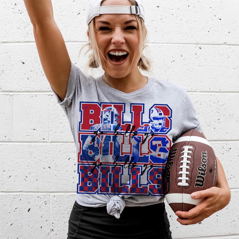 May include: A gray t-shirt with a Bills graphic in red, white, and blue, paired with a white baseball cap and a brown football. The shirt features the word "Bills" repeated multiple times with player images.
